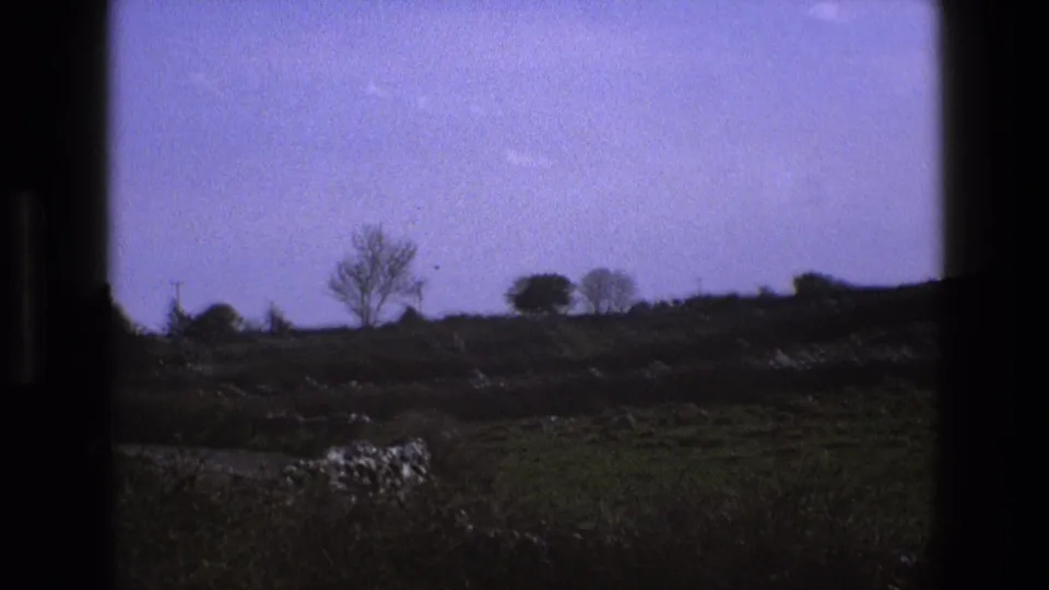 1969 England 8mm frame showing a shadowed hillside, bare deciduous trees, low stone walls, and a lilac-blue sky.
