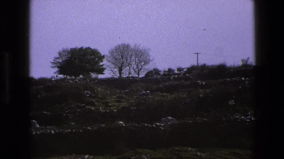 1969 England 8mm frame of a rocky rural hillside with scattered trees, dark foreground, and a distant utility pole.