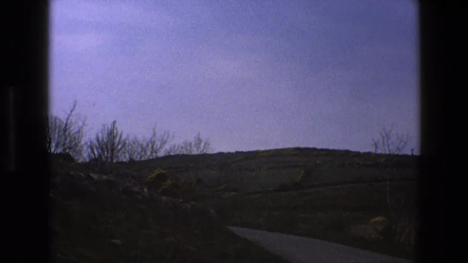1969 8mm view of an English rural road curving through rolling fields, framed by dark window edges and bare roadside trees.