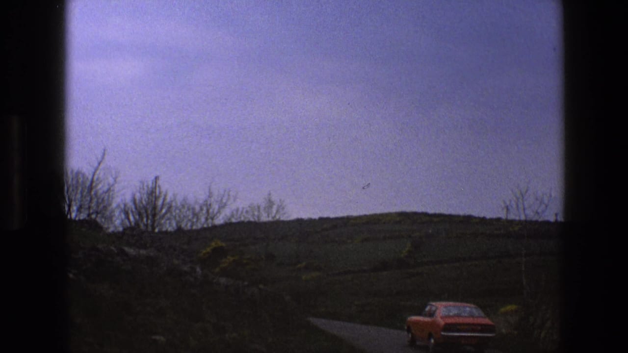 Red car driving away on a narrow rural road beneath rolling hills.