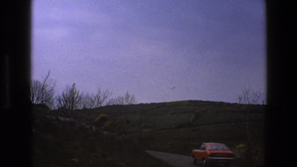 Red car travels away along a narrow English countryside road in 1969, with stone walls, bare trees, and green hills in the background.