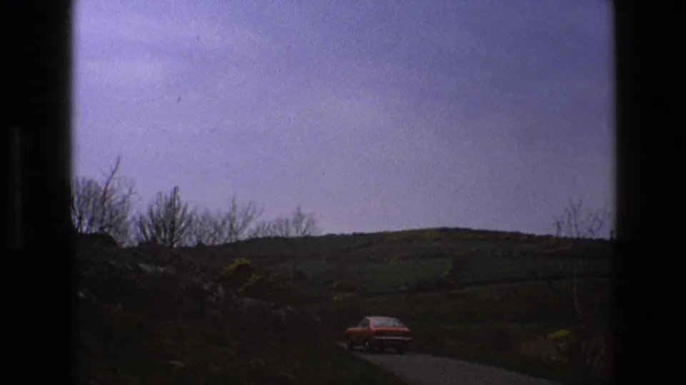 The red car continues away from the camera on a rural English road, partly framed by dark film or window edges and open fields.