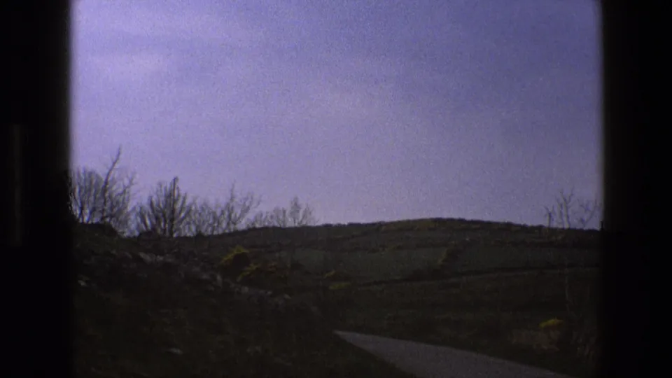 Empty countryside road in England after the car has passed, with rolling hills, roadside brush, and a pale purple-blue sky on 8mm film.
