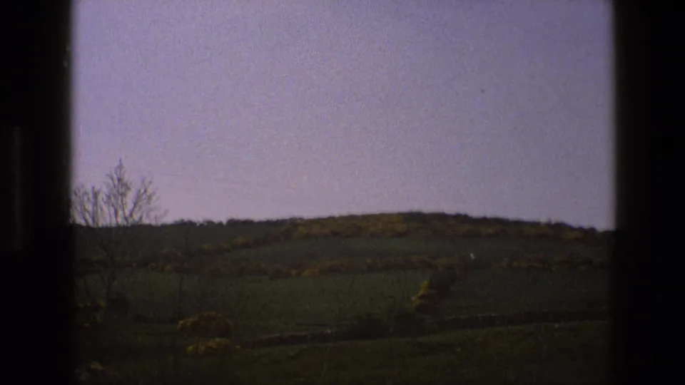End frame from a 1969 England home movie showing open countryside, low stone walling, yellow bushes, and a pale sky.
