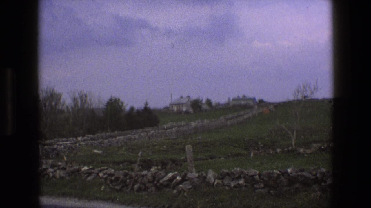 1969 England countryside frame with distant hilltop houses, dry stone walls, green fields, leafless trees, and cloud cover.