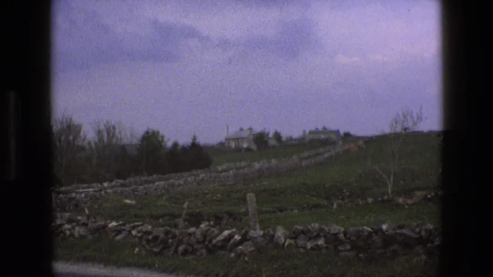 1969 England countryside frame with distant hilltop houses, dry stone walls, green fields, leafless trees, and cloud cover.
