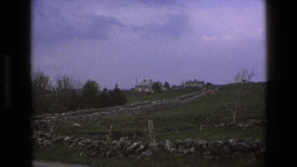1969 England 8mm view of stone-walled fields leading toward distant rural houses under a soft cloudy sky.