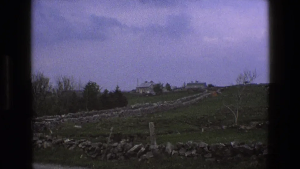 1969 England countryside frame with foreground stone wall, open fields, distant buildings, bare trees, and purple-tinged clouds.