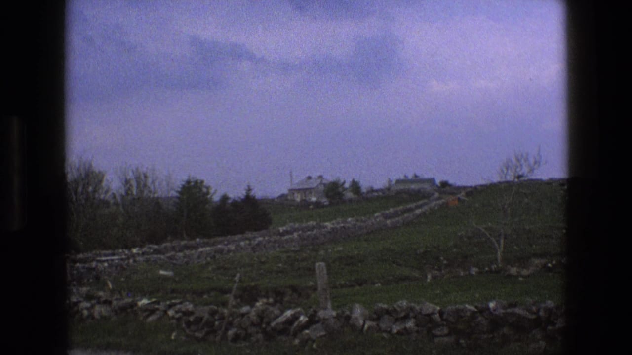 Distant stone farmhouse beyond green fields and dry-stone walls under a cloudy sky.