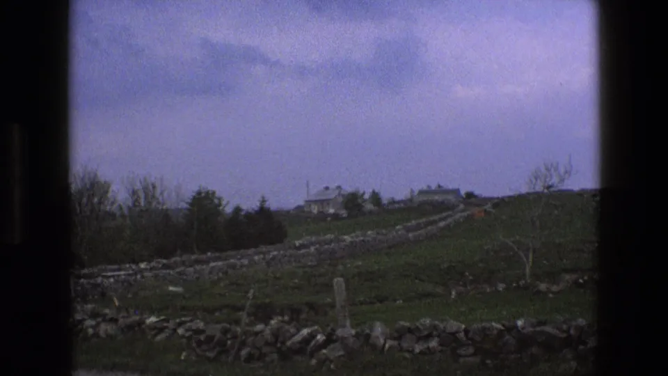 1969 England 8mm frame showing a distant stone farmhouse, green pasture, and dry-stone wall lines seen through a dark window edge.