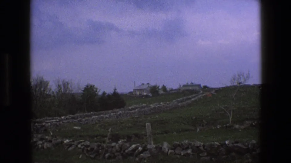1969 England countryside view with stone wall lines crossing pasture toward farmhouse buildings below a muted blue-gray sky.