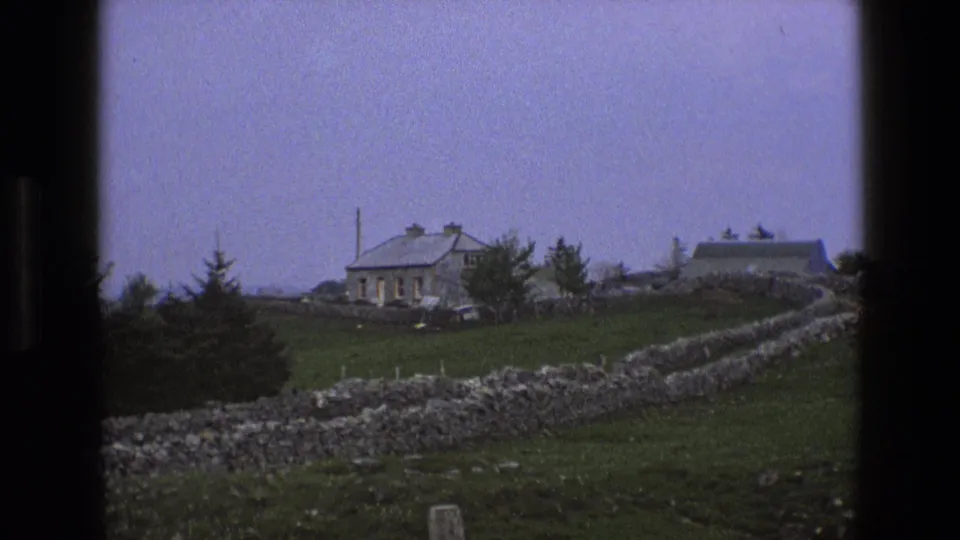 1969 England rural frame showing a stone farmhouse and outbuilding behind a curving dry-stone wall, with open pasture and pale sky.