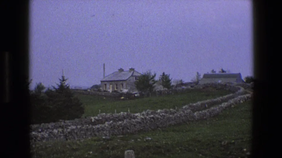 1969 England 8mm frame showing a stone cottage, gray roof, pasture, and dry-stone walls viewed through dark frame edges.