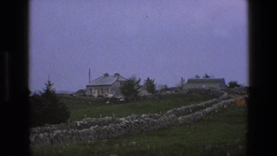 1969 England home-movie frame with a stone cottage, distant farm building, dry-stone wall, grass, and pale sky.