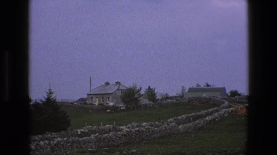 1969 England 8mm view of a stone cottage and dry-stone field boundary, with trees and farmland in soft focus.