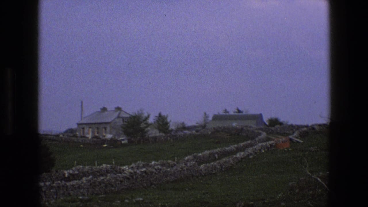 1969 England home-movie frame showing dry-stone walls, grass, a distant house, and a pale outbuilding beyond a winding path.