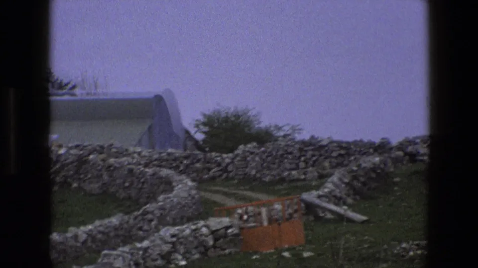 1969 England rural frame with dry-stone walls, a curved-roof outbuilding, grass, trees, and a small orange gate or barrier.