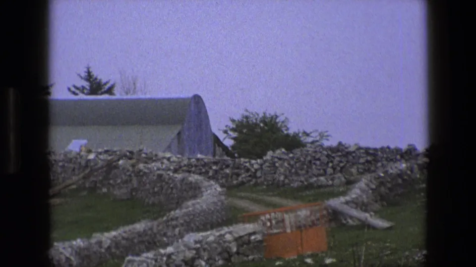 1969 England home-movie view across dry-stone walls toward a pale outbuilding, with grass, trees, and an orange barrier in the foreground.