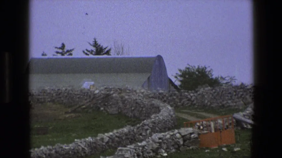 1969 England frame showing a grassy rural enclosure, dry-stone walls, a pale curved-roof building, trees, and an orange barrier.