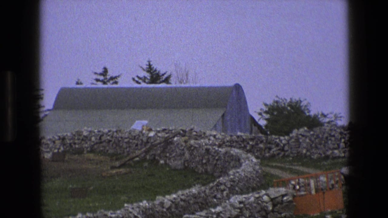 1969 rural England farm view with an arched-roof building, dry-stone wall, grassy field, and pale sky within dark 8mm frame edges.