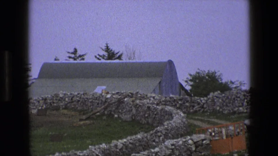1969 rural England farm view with an arched-roof building, dry-stone wall, grassy field, and pale sky within dark 8mm frame edges.