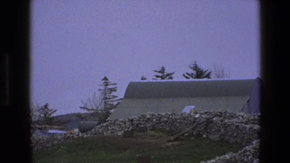 1969 8mm frame of an English farm with a curved-roof outbuilding, dry-stone wall, grassy field, and trees under a pale sky.