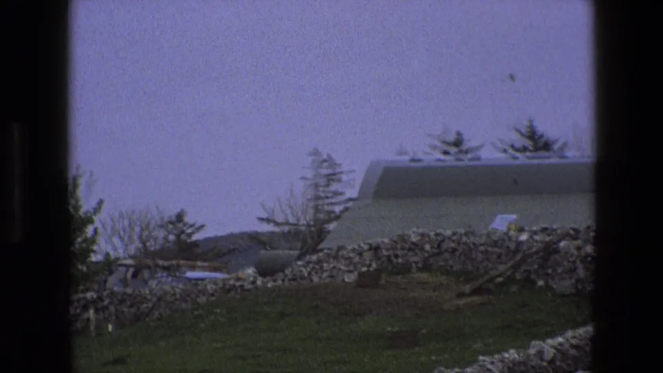 Final 1969 rural England frame with the barn partly cropped, dry-stone wall in front, grassy field below, and trees against a pale sky.