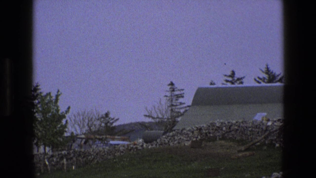 Stone house roofline behind trees and a dry-stone wall, seen through dark film-frame edges.
