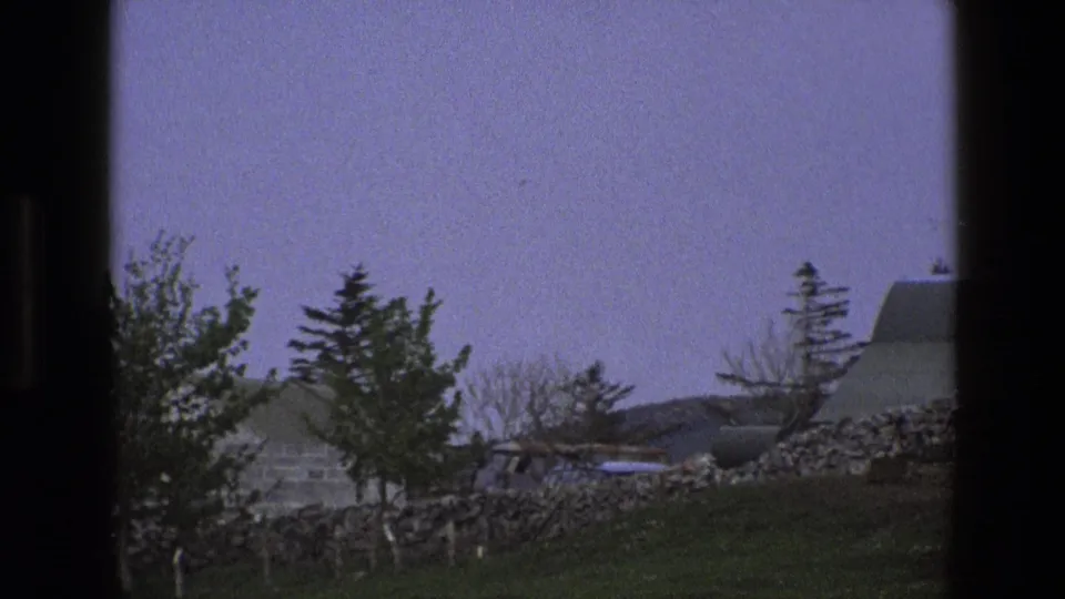 1969 England home-movie frame with trees, a dry-stone wall, and a low stone building partly obscured by the handheld framing.