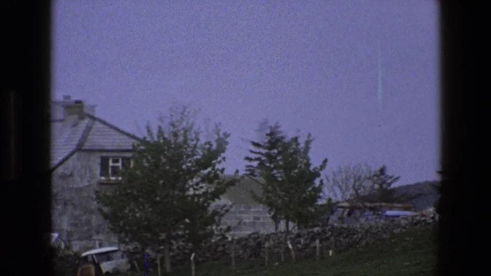 1969 England 8mm frame showing a stone house with slate roof, chimneys, trees, grass, and a dry-stone wall.