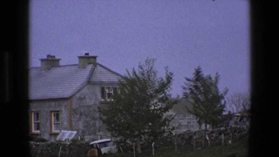 1969 England home-movie frame showing a stone house facade, slate roof, chimneys, trees, and a dry-stone garden wall.