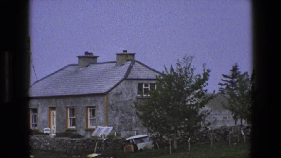 1969 England 8mm frame of a rural stone cottage behind a low wall, seen through dark vertical framing.
