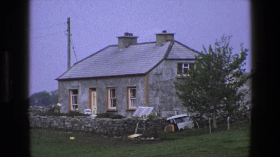 1969 England 8mm frame showing a stone cottage, yard wall, small van, leaning panel, and trees in a rural setting.