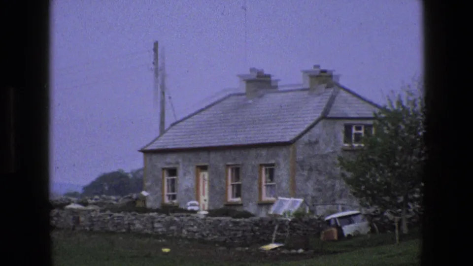 1969 England 8mm frame of a rural stone cottage viewed closer, with a low wall, grass, and small van nearby.