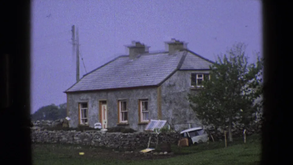 1969 England 8mm frame showing the rural cottage, yard wall, small van, leaning panel, and surrounding trees.