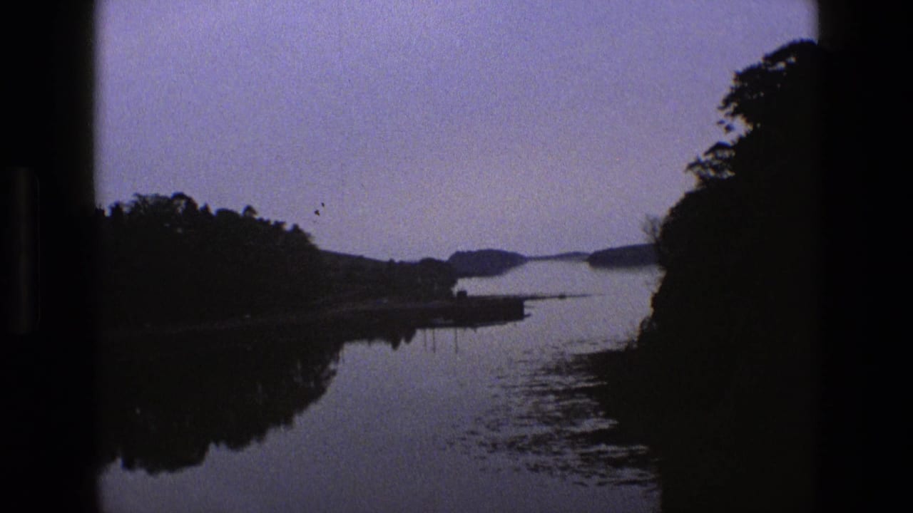 1969 England 8mm frame showing a calm river or inlet at dusk, with silhouetted trees, sky reflection, distant low land, and dark film borders.