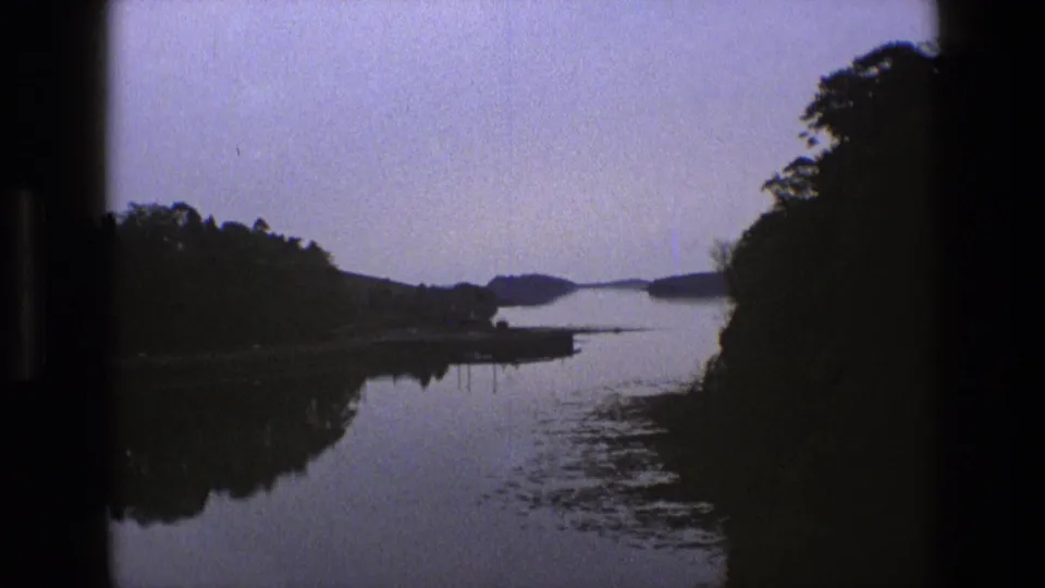 1969 England 8mm frame of a quiet waterway viewed from a moving position, with dark trees, reflective water, and low distant shoreline.