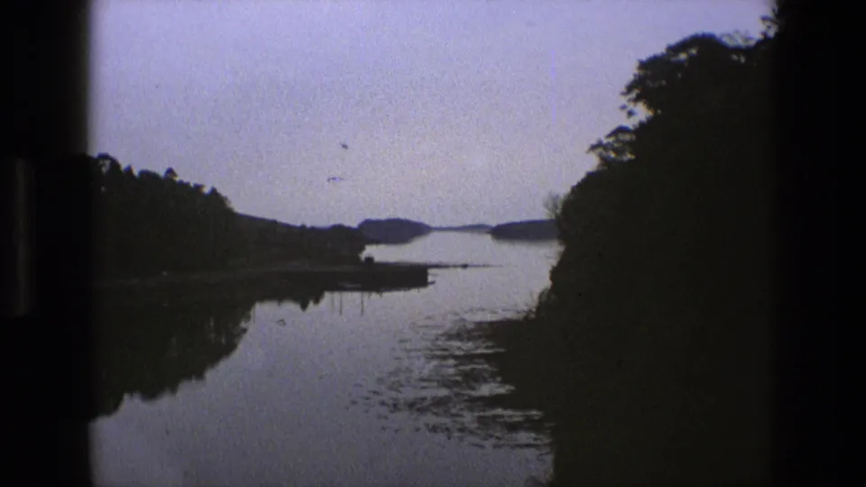 1969 England 8mm frame with dark wooded banks, a small jetty or platform, and muted sky reflected in a calm river surface.