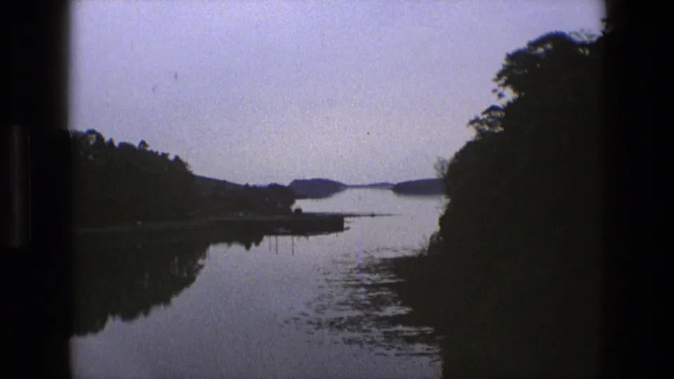 1969 England 8mm frame showing a tranquil river view at dusk, with tree silhouettes on both sides and a soft reflection across the water.