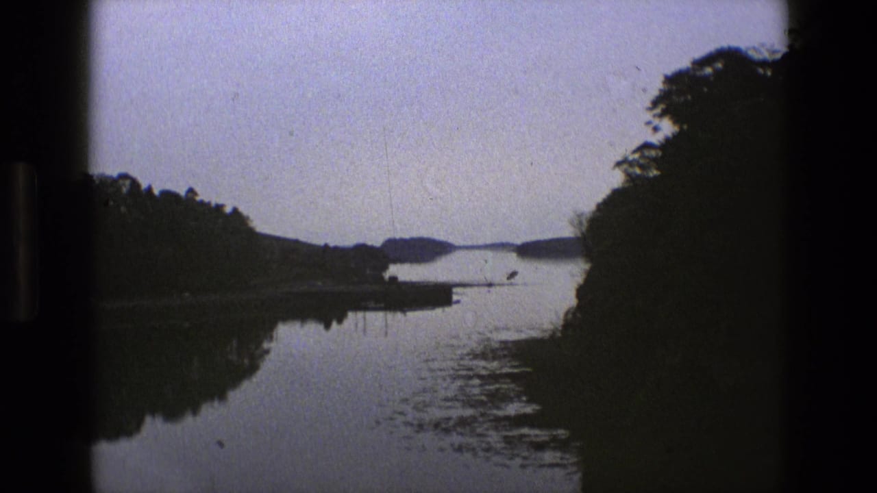 1969 England 8mm frame showing a calm lake, tree-lined banks, distant hills, and dark film borders from a moving viewpoint.