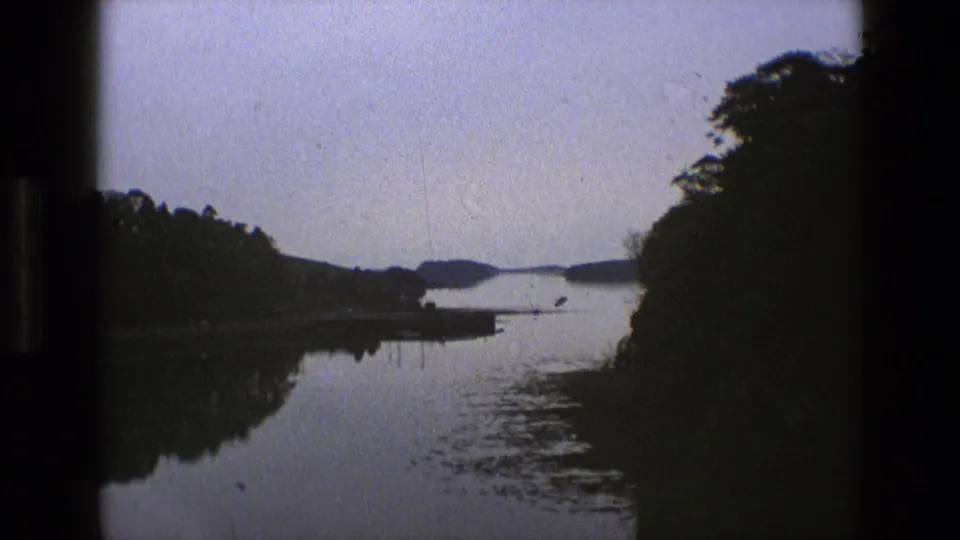 1969 England 8mm frame showing a calm lake, tree-lined banks, distant hills, and dark film borders from a moving viewpoint.