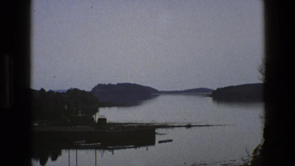 1969 England 8mm frame with a broad lake, small lakeside landing, moored boats, wooded edges, and low hills beyond.