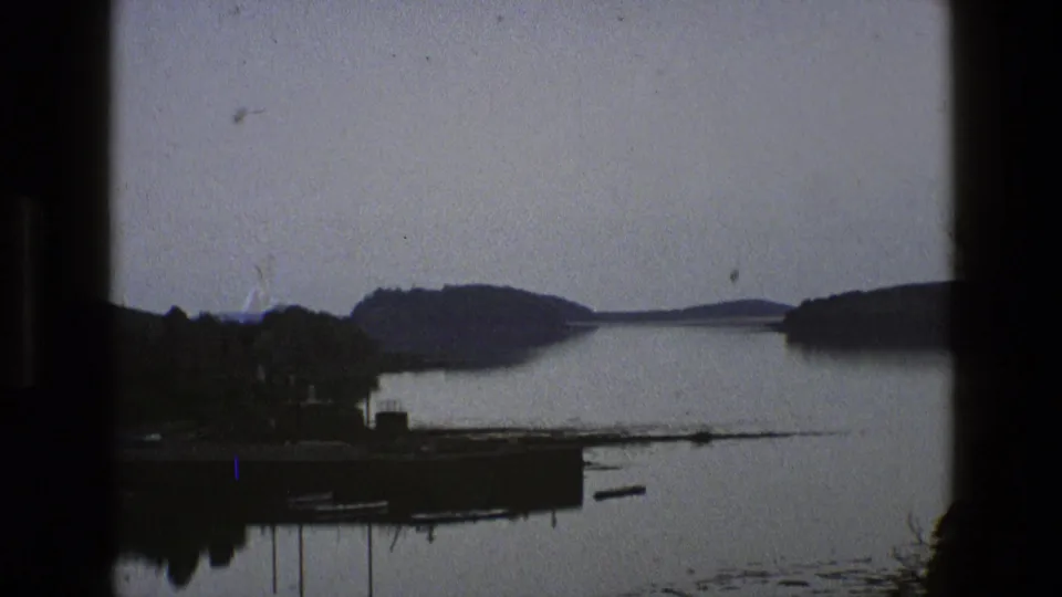 1969 England 8mm frame showing calm water, a shoreline landing, small boats, tree cover, and low hills under grey light.