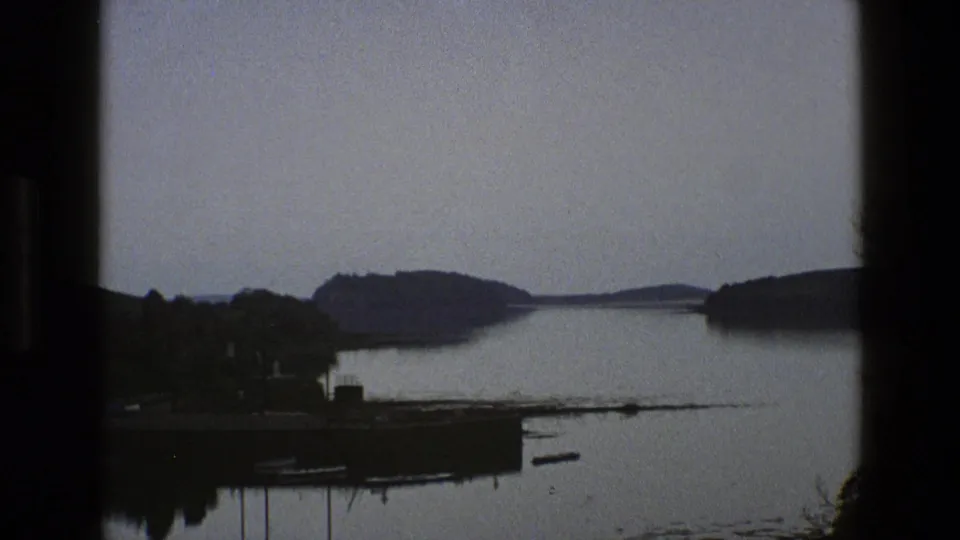 1969 England 8mm frame of a quiet lake with wooded banks, a small landing, boats, distant hills, and visible film grain.