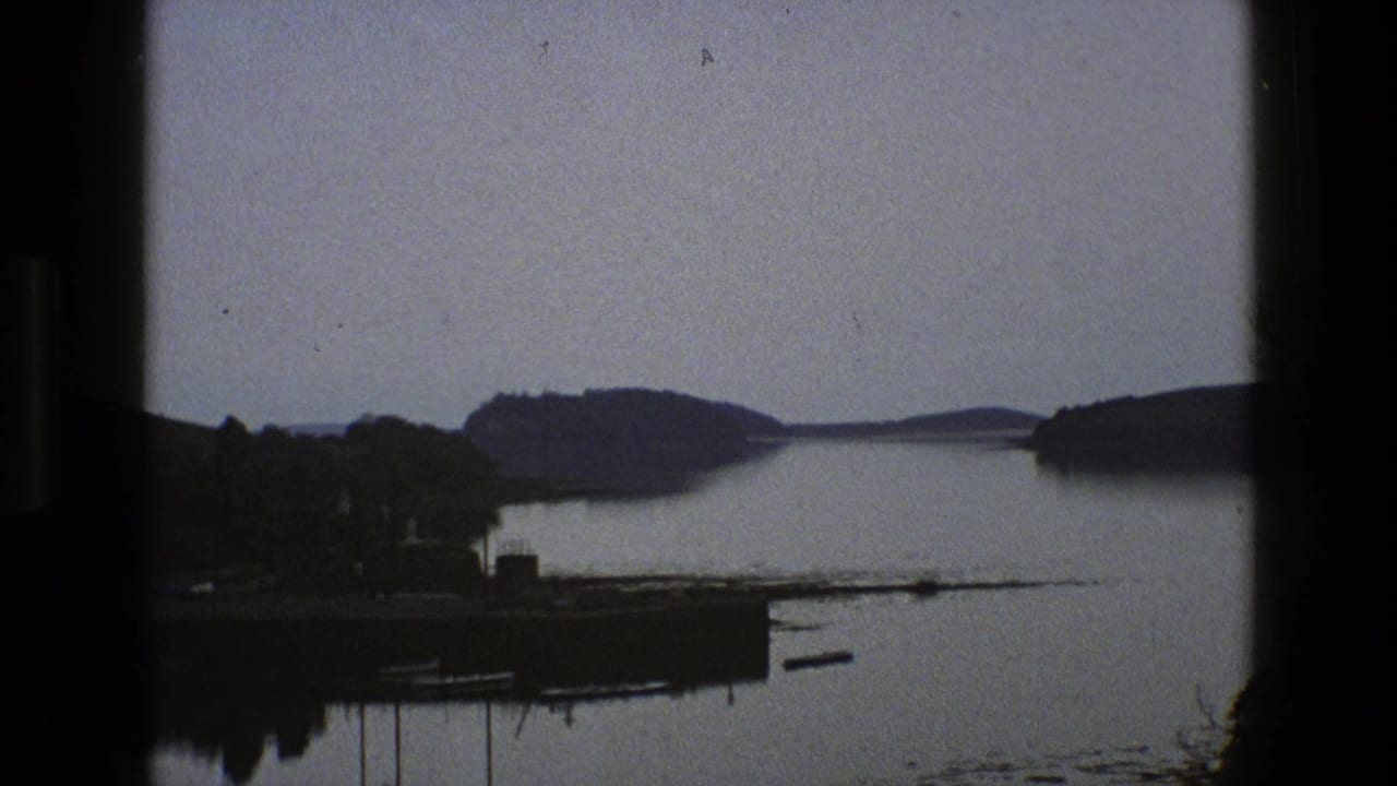1969 England 8mm frame showing calm water, distant wooded hills, a small shoreline structure and dark rounded frame edges.