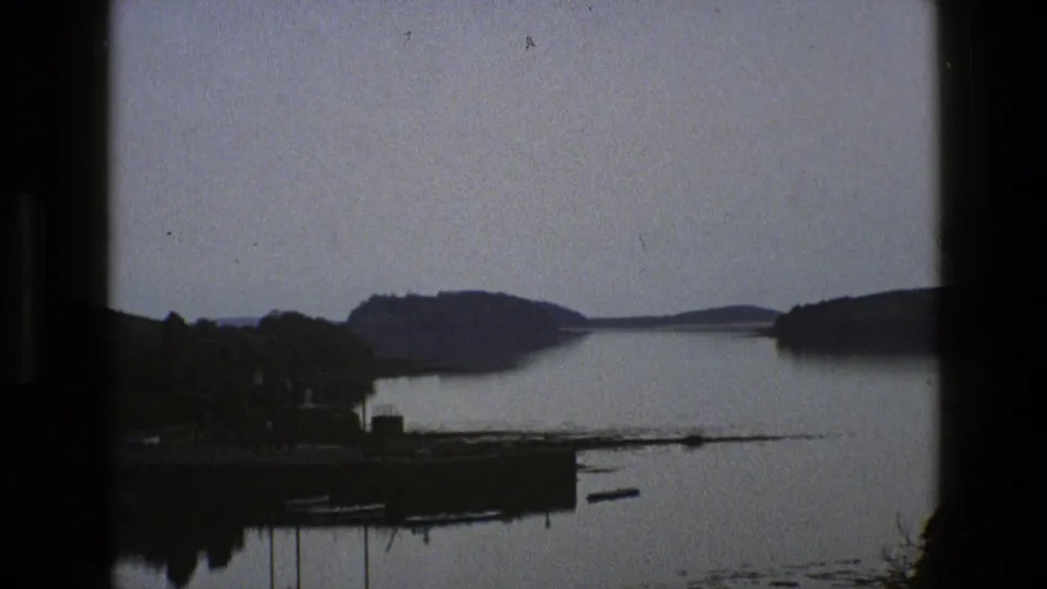 1969 England 8mm frame showing calm water, distant wooded hills, a small shoreline structure and dark rounded frame edges.