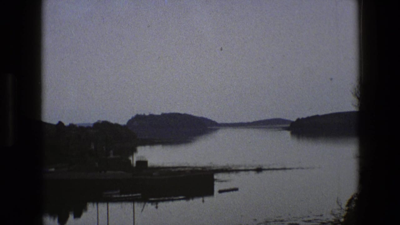 Wide view of still water, dark wooded shoreline and distant low hills.
