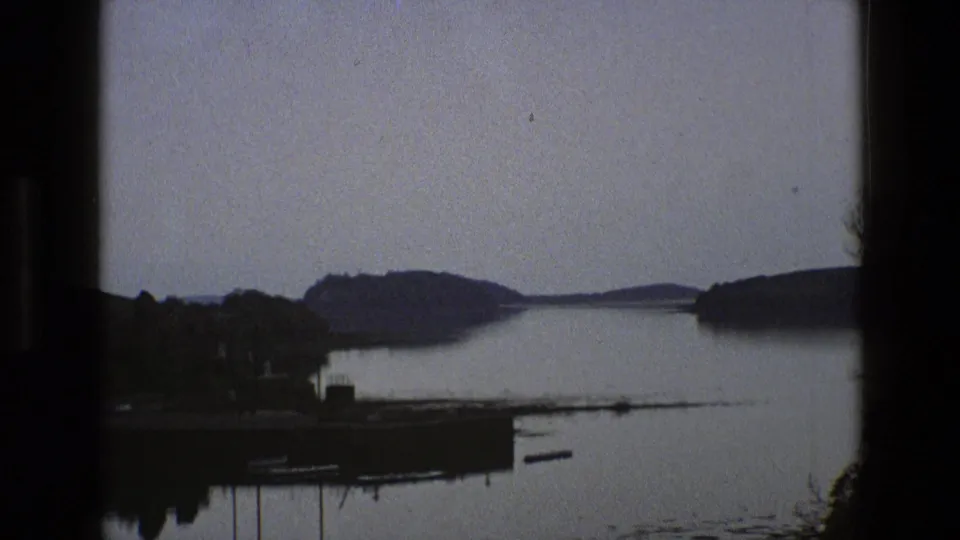 1969 England 8mm frame with still water, tree-covered shoreline, distant hills and soft overcast light.