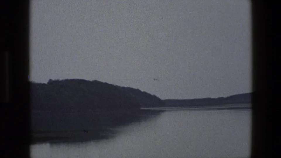 1969 England 8mm frame showing a wooded hill descending toward calm water beneath a pale overcast sky.