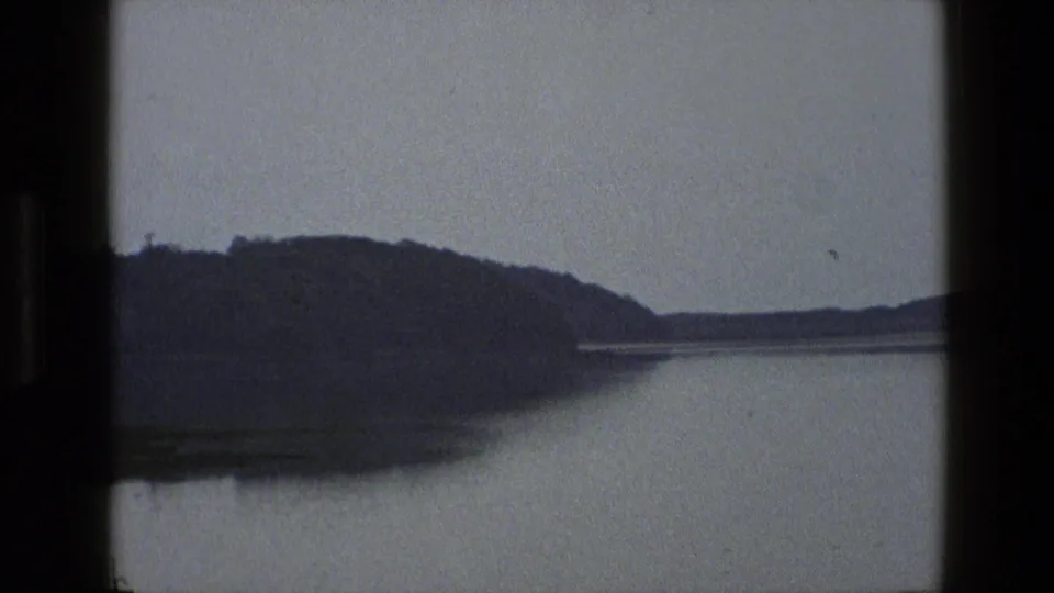 1969 England 8mm frame showing a tree-covered headland, smooth water and a distant low shoreline.